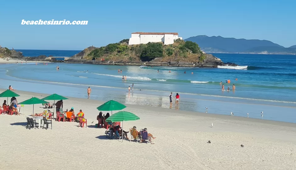 Wide view of Praia do Forte in Cabo Frio during the low season, with crystal-clear sea, blue skies, few people on the white sand, and the São Mateus Fort in the background.