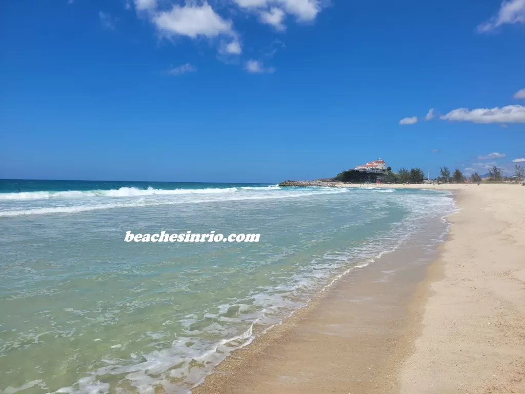 Crystal-clear waters and perfect waves at Praia de Itaúna on a sunny summer morning, with white sandy shores and the Igreja de Nossa Senhora de Nazareth in the background.