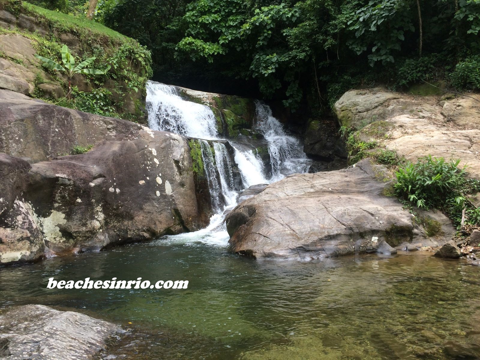Natural pool formed at the base of a waterfall in the Atlantic Forest, with clear flowing water surrounded by rocks and lush green tropical vegetation.